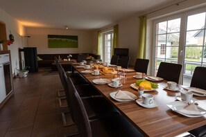 Ein Essbereich mit einem langen Tisch, gedeckt für das Frühstück, Stühlen und Blick ins Freie durch große Fenster.