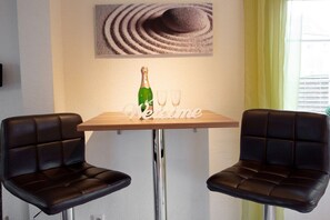 Ein kleiner Tisch mit einer Champagnerflasche und zwei Gläsern, zwei schwarze Stühle und ein Kunstwerk über dem Tisch.