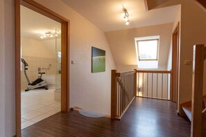 Ein Zimmer mit Holzboden, einer Treppe mit Geländer, einem Fenster und einer Tür zu einem Badezimmer mit Dusche und Ergometer.