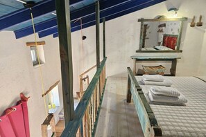 A room with a wooden staircase, a bed with white bedding, a small window, and a mirror above the bed. - Studio Komos Gallery (Pitsidia)