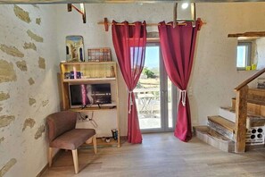 A room with a wooden bench, a television, a shelf with decorative items, and a view through a glass door. - Studio Komos Gallery (Pitsidia)