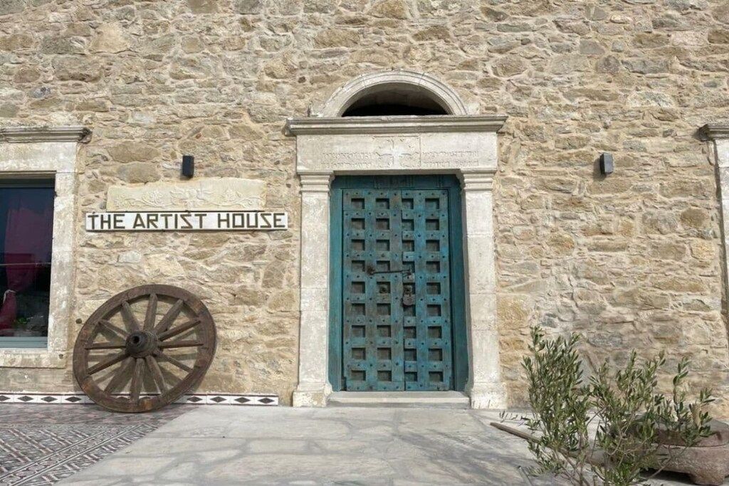 A stone building with a sign reading 'THE ARTIST HOUSE' and a large wooden wheel to the left.