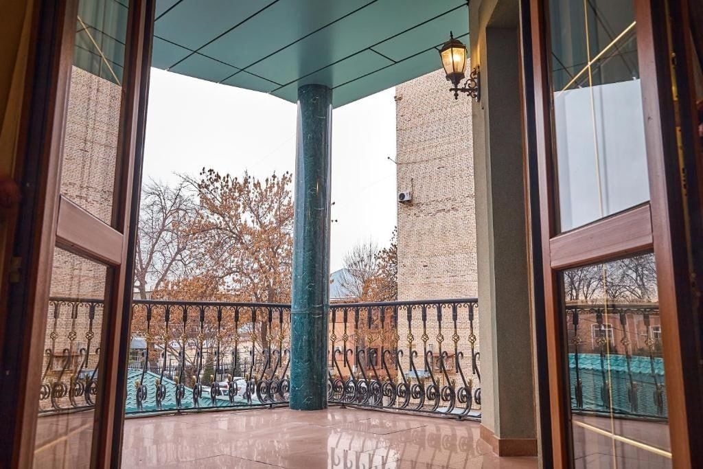 Un espacio al aire libre cubierto con barandilla metálica, un pilar central y vistas a un edificio de ladrillo y árboles.