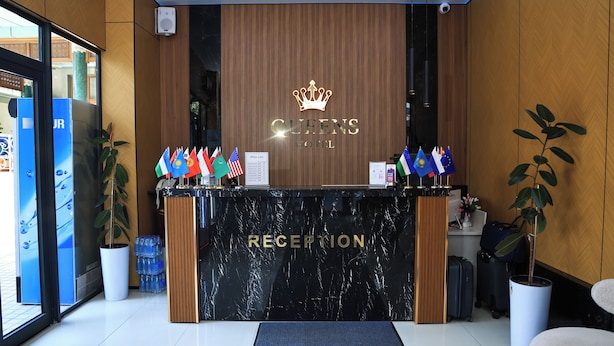 A hotel reception desk with a 'RECEPTION' sign, multiple flags, a 'Price List' sign, and a crown logo above the name 'QUEENS HOTEL'. - Queens Hotel (Tashkent)