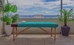 Ett massagebord med ett turkosfärgat överdrag, placerat på en balkong med krukväxter och utsikt över havet.