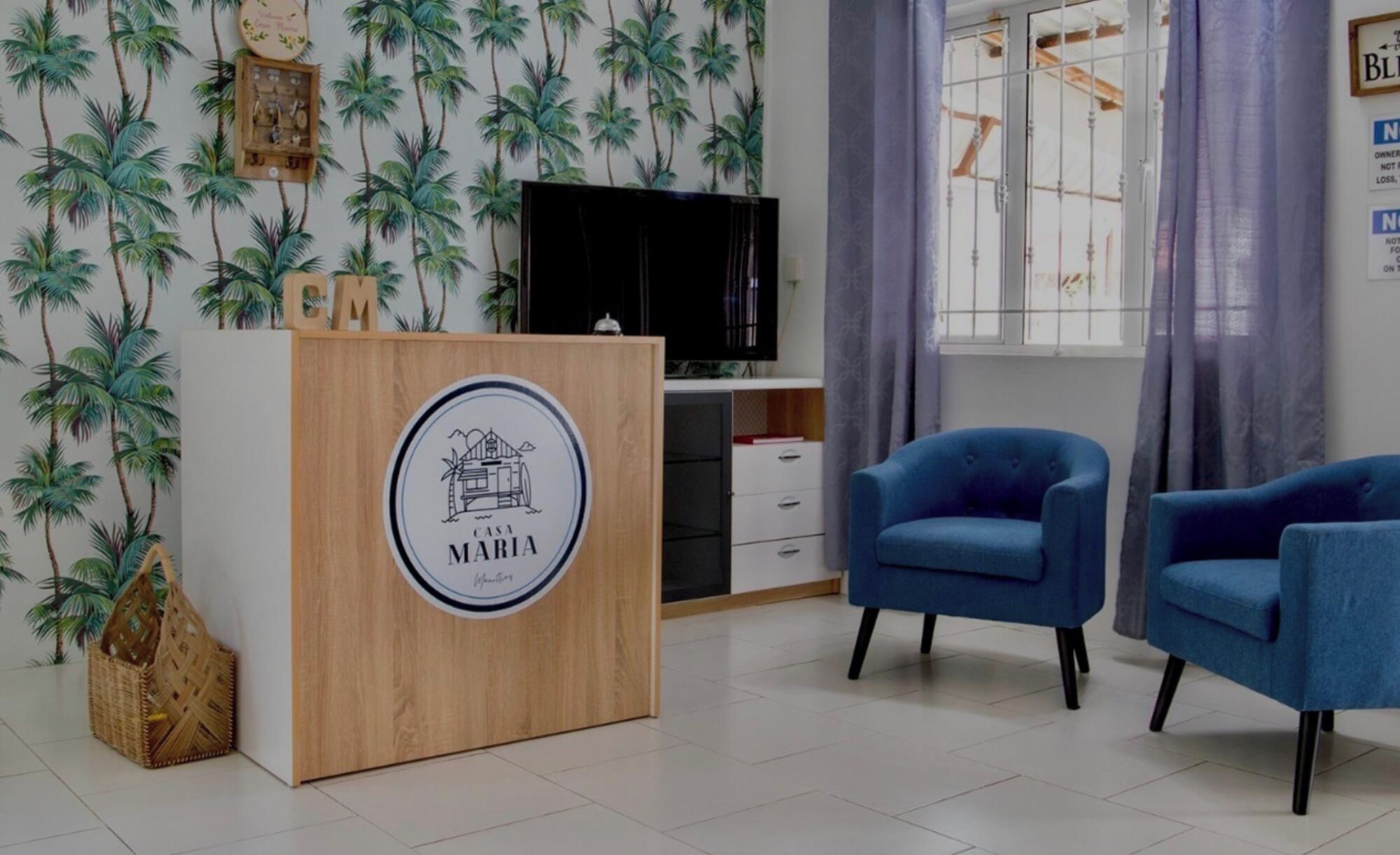Une pièce avec un comptoir d’accueil en bois, deux fauteuils bleus, un téléviseur et une fenêtre avec des rideaux.