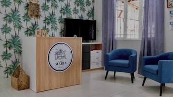 Une pièce avec un comptoir d’accueil en bois, deux fauteuils bleus, un téléviseur et une fenêtre avec des rideaux.