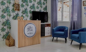 A room with a wooden reception desk, two blue armchairs, a television, and a window with curtains.