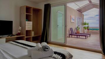 A hotel room with a bed, a TV, a wardrobe, a balcony with a view of the sea, and a seating area with plants.