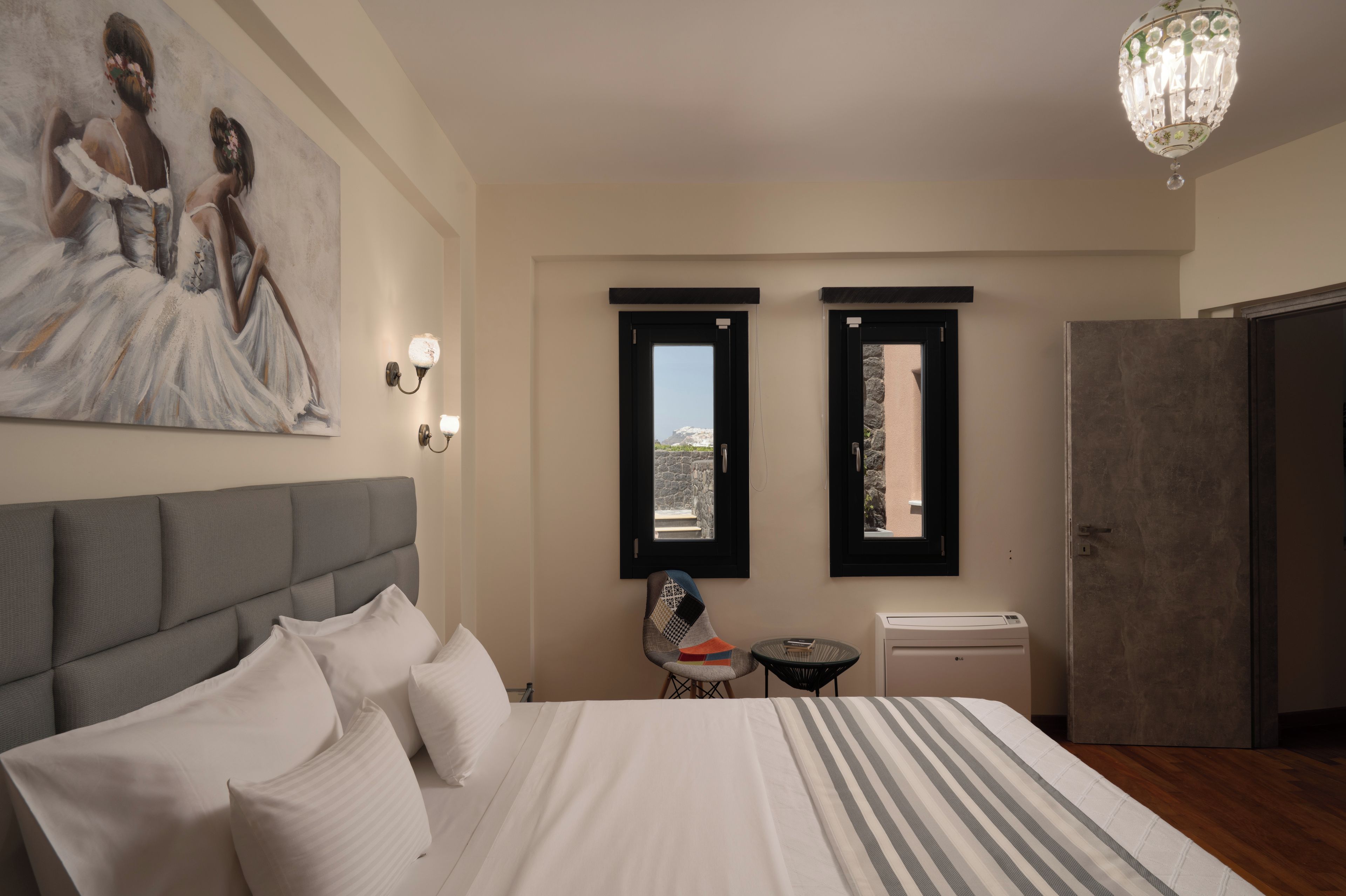 A hotel room with a bed, a painting on the wall, two windows with black frames, a chandelier, and a small table with a chair.