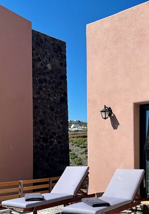 A rooftop terrace with two lounge chairs, a stone wall, and a building with a textured facade.