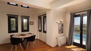 Ein modernes Wohnzimmer mit Holzboden, einem runden Esstisch mit Stühlen, einer integrierten Klimaanlage und einem großen Schiebeglasfenster mit Blick ins Freie.