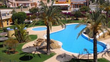 Un resort con una amplia piscina, palmeras y un área cercada con sombrillas.