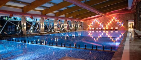 Una amplia piscina interior con suelo de baldosas, rodeada de sillones de descanso y una estructura de techo de madera.