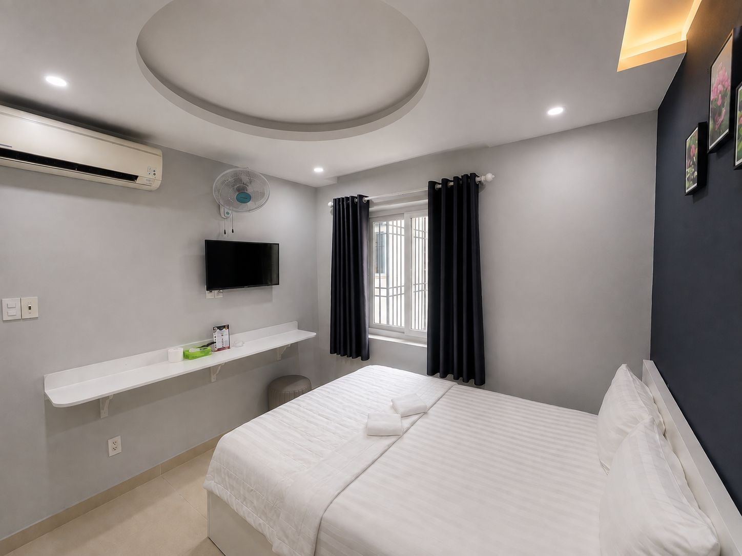 A hotel room with a bed, a TV, a fan, a window with curtains, and a wall-mounted shelf.