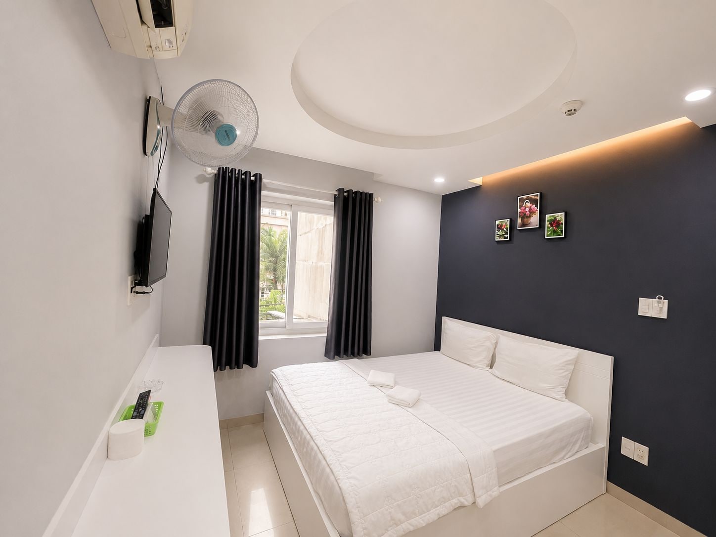 Un dormitorio moderno con cama, mesita de noche, televisor, ventana con cortinas y una pared decorada con cuadros.