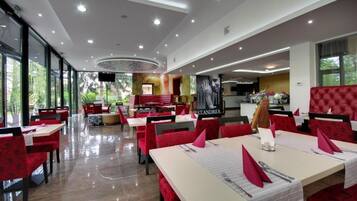A restaurant with red upholstered chairs and tables, a modern ceiling design, and large windows allowing natural light.