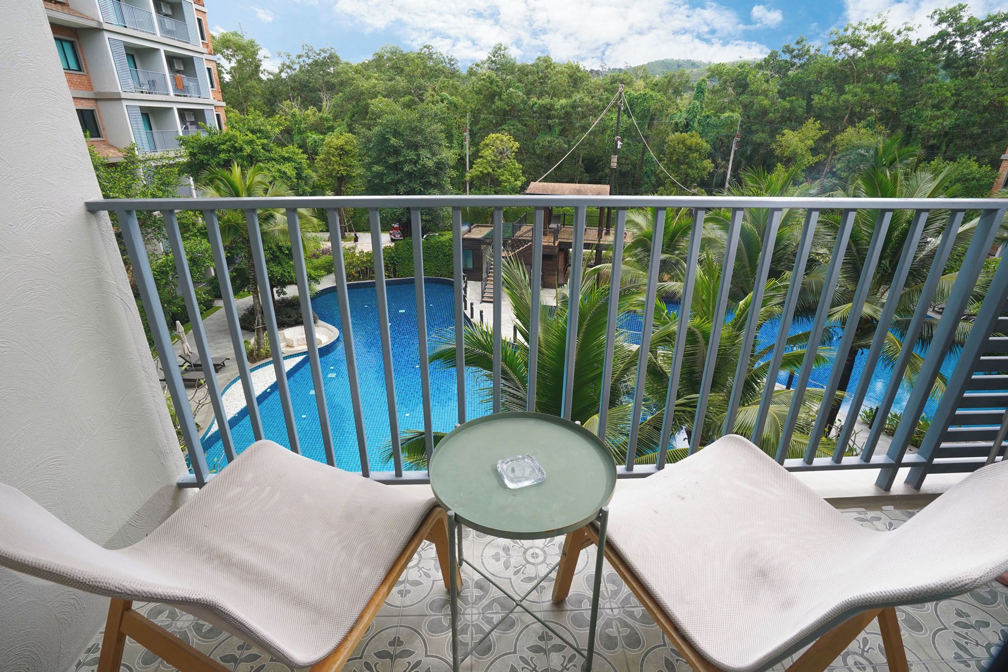 Un balcon avec deux fauteuils blancs dotés de coussins et une petite table, donnant sur une piscine circulaire entourée d’une végétation luxuriante et sur un immeuble à plusieurs étages.