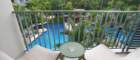 Un balcon avec deux fauteuils blancs dotés de coussins et une petite table, donnant sur une piscine circulaire entourée d’une végétation luxuriante et sur un immeuble à plusieurs étages.