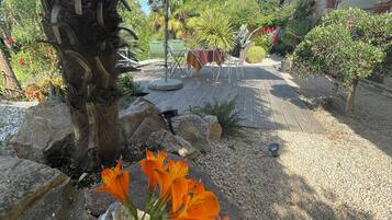 Un espacio al aire libre con mesa y sillas bajo una pérgola, rodeado de rocas y plantas.