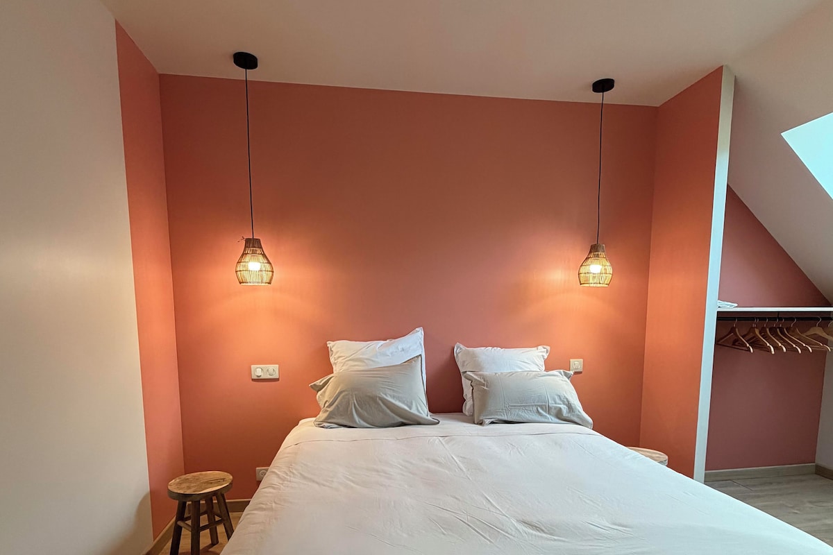 A bedroom with a bed, two hanging lights, a stool, and a closet with hanging clothes.