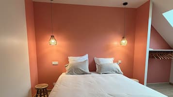 Un dormitorio con una cama, dos lámparas colgantes, un taburete y un armario con ropa colgada.