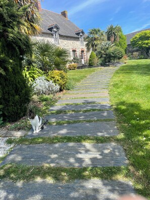 Ein gepflasterter Weg führt zu einem Steinhaus mit geneigtem Dach, umgeben von üppiger Vegetation. Eine Katze sitzt auf dem Weg.