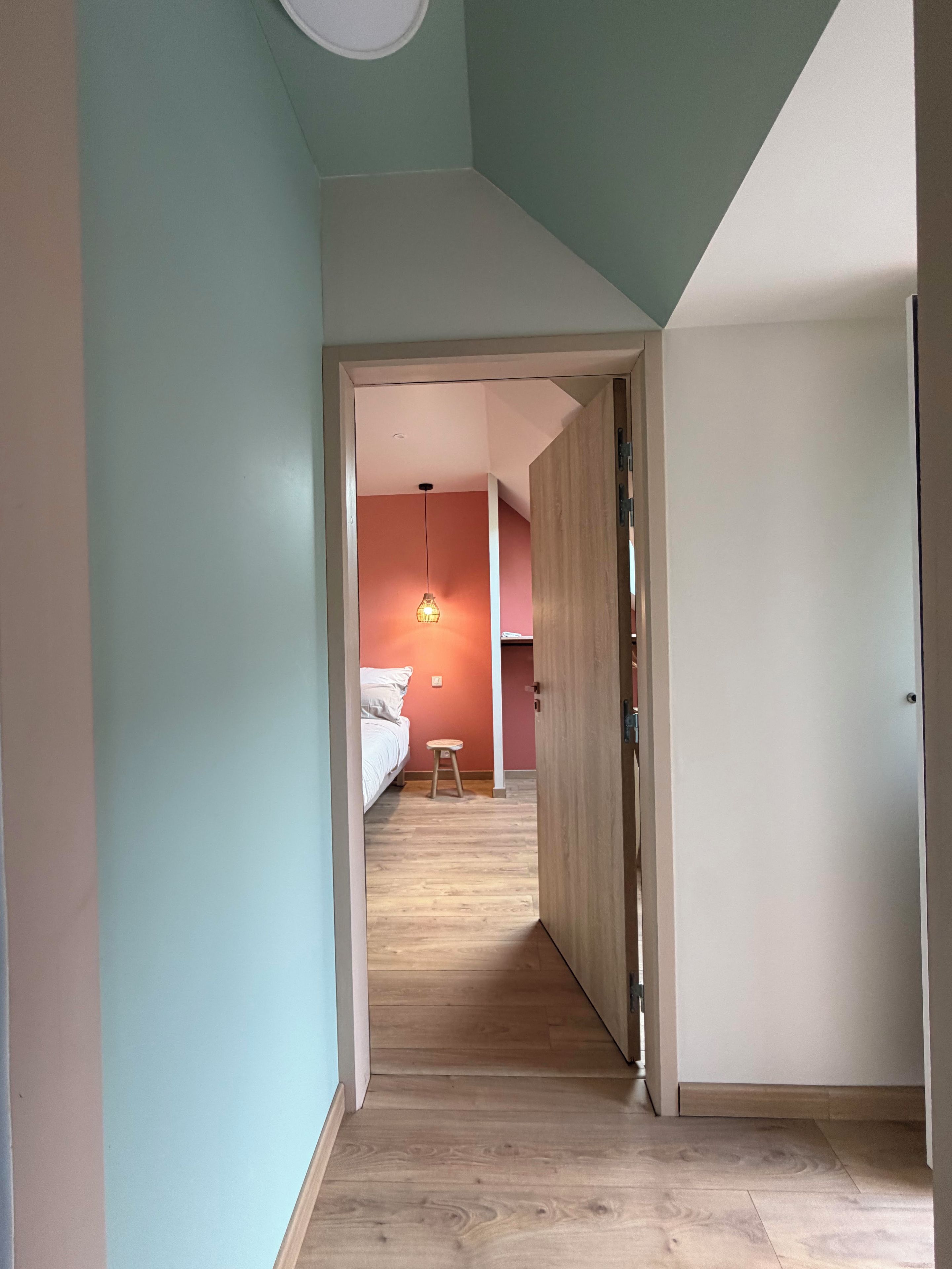 Un couloir étroit mène à une chambre qui contient un lit et un tabouret, le sol est en bois et une porte se trouve sur le côté droit.