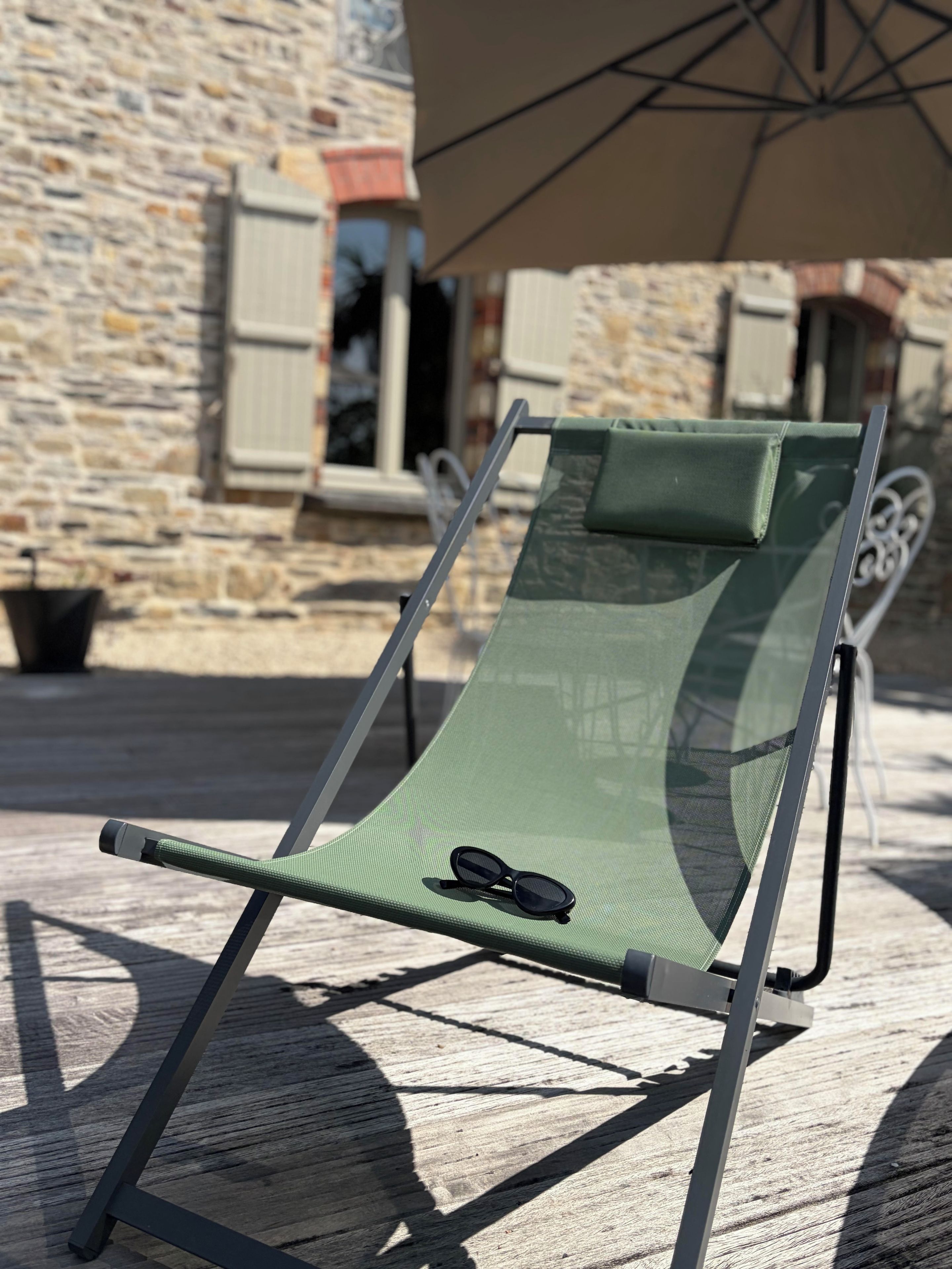 A green lounge chair with a pair of sunglasses on it, positioned on a wooden deck with a stone wall in the background.