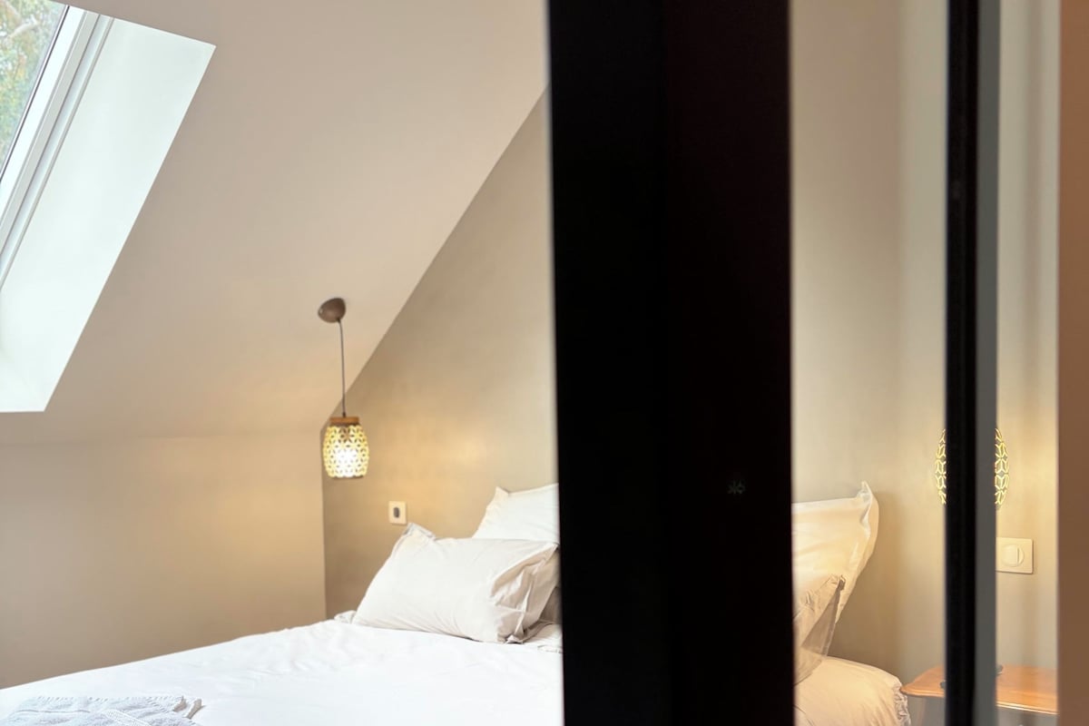 A bedroom with a bed, pillows, a bedside table, a hanging light fixture, and a skylight.