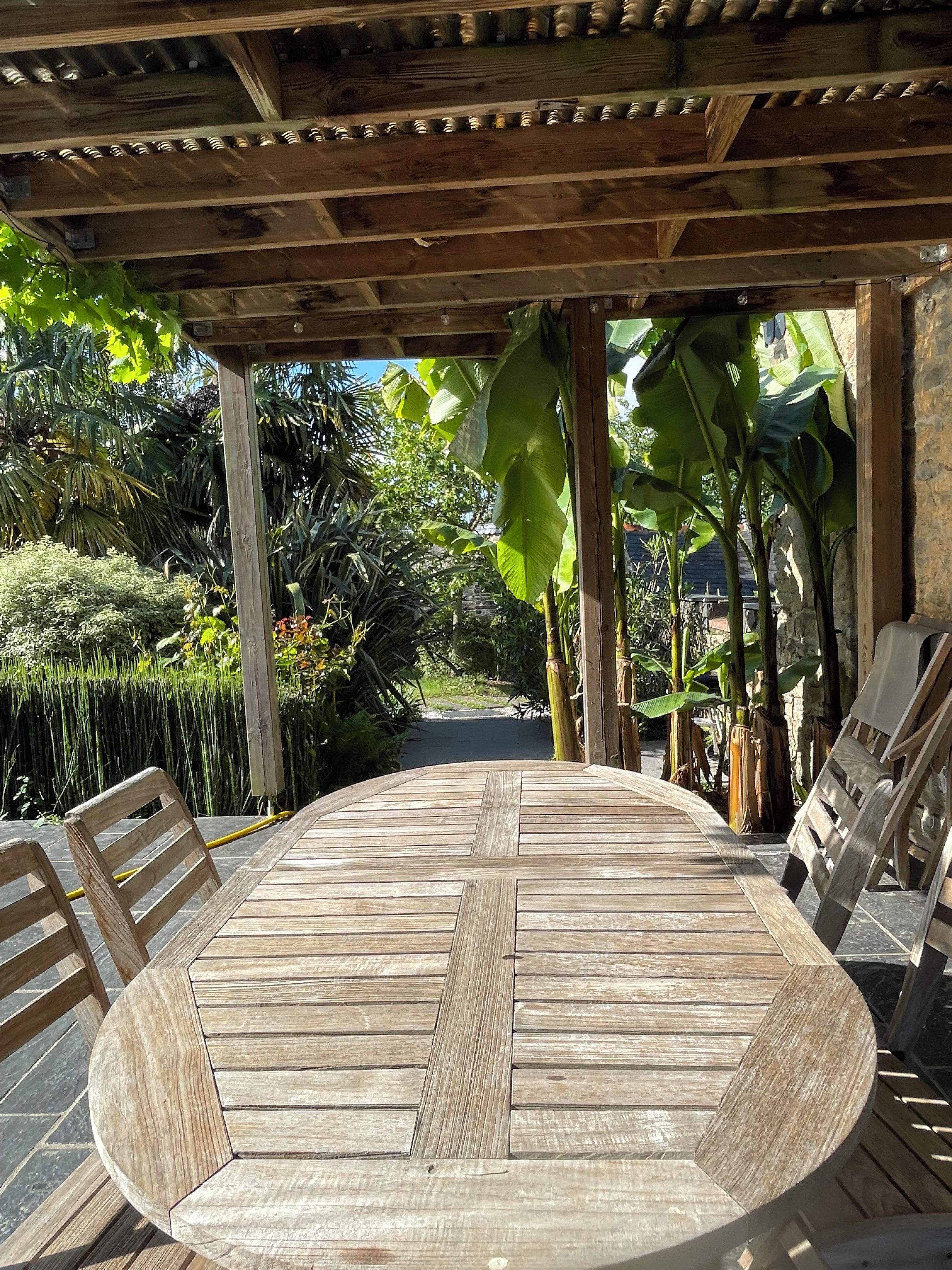 Una mesa de madera redonda con sillas bajo una pérgola de madera, rodeada de exuberante follaje tropical.
