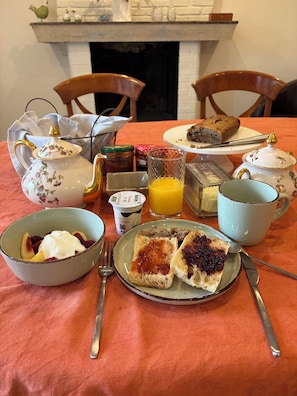 Un desayuno con tetera, tazas, un plato de tostadas y mermelada, un vaso de zumo de naranja y una hogaza de pan sobre una mesa.
