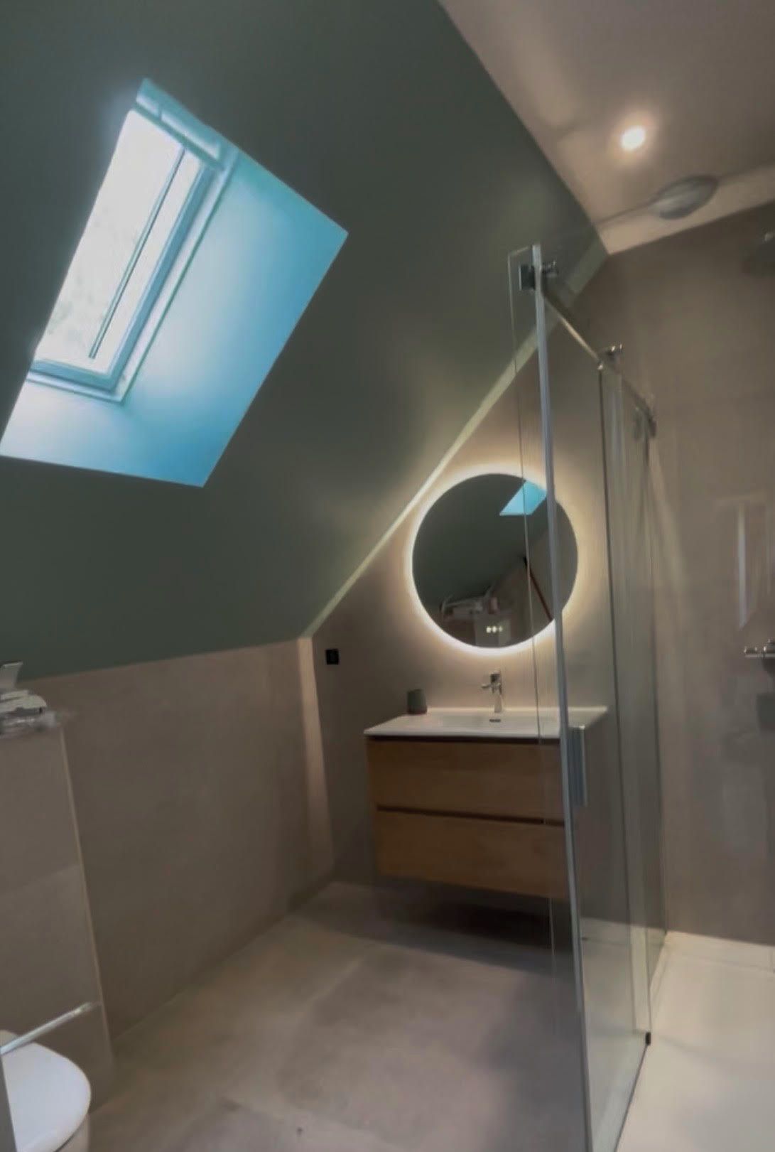 Un baño moderno con tragaluz, un espejo redondo y cabina de ducha de vidrio.