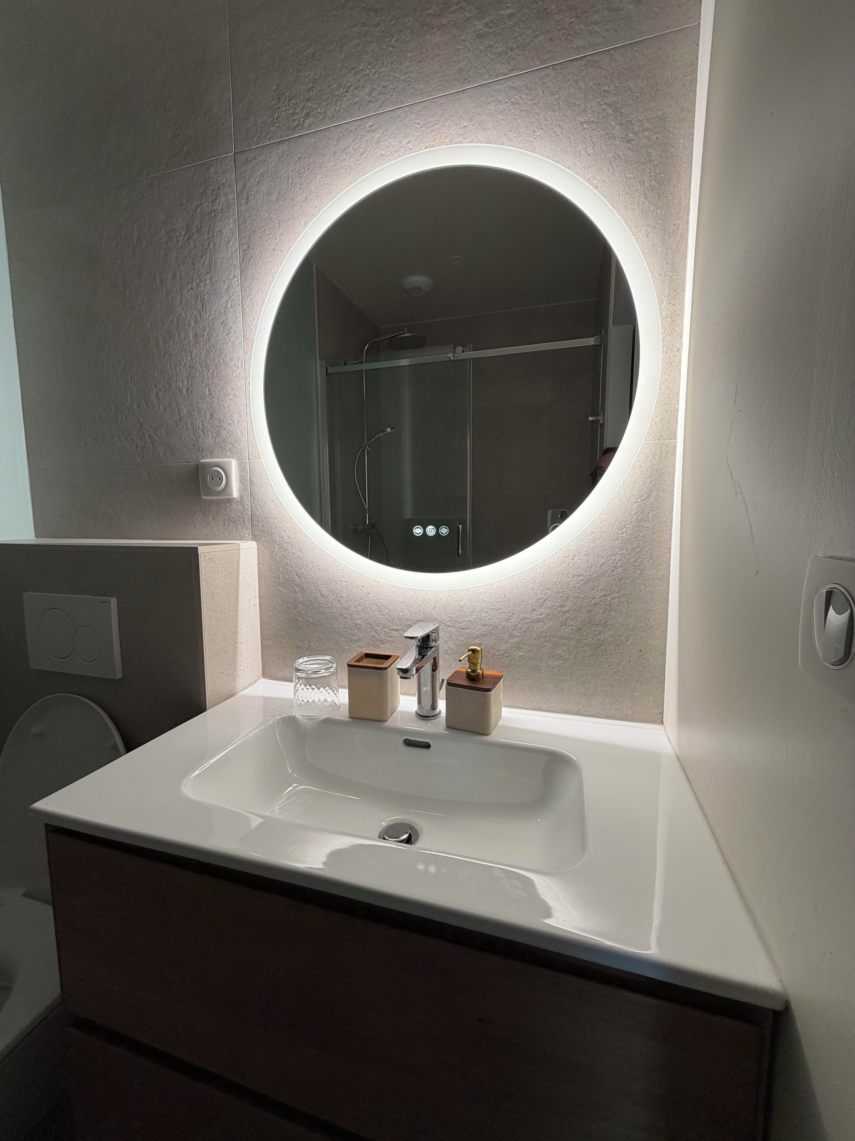 A bathroom with a round mirror, a white sink, and a shower visible in the background.