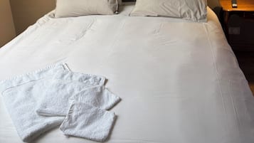 Ein ordentlich bezogenes Bett mit weißen Leinen und Kissen, zwei Pendelleuchten mit gemusterten Lampenschirmen und ein Nachttisch aus Holz mit einer Fernbedienung.