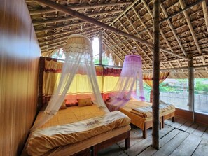 Quarto com duas camas, cada uma com colcha de dossel e mosquiteiro. O quarto possui piso de madeira e telhado de palha.