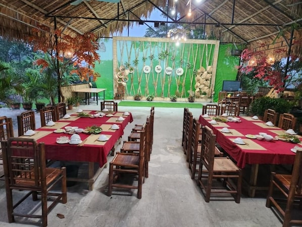 Un comedor interior con mesas dispuestas para una comida, un escenario con un telón de fondo y plantas tropicales.