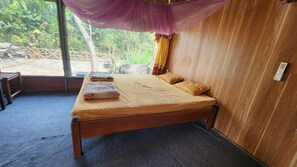 Uma cama de solteiro com estrutura de madeira, um edredom bege e duas almofadas. O quarto possui uma parede de madeira e uma janela com vista para o verde lá fora.