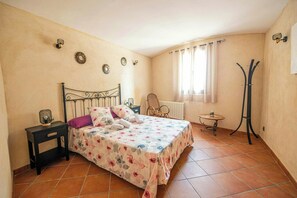 Un dormitorio con cama, mesita de noche, silla, radiador, ventana con cortinas y perchero.