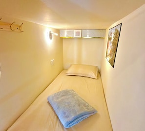 Une petite chambre avec un lit simple, un oreiller, une serviette pliée, une lampe fixée au mur et une prise de courant.