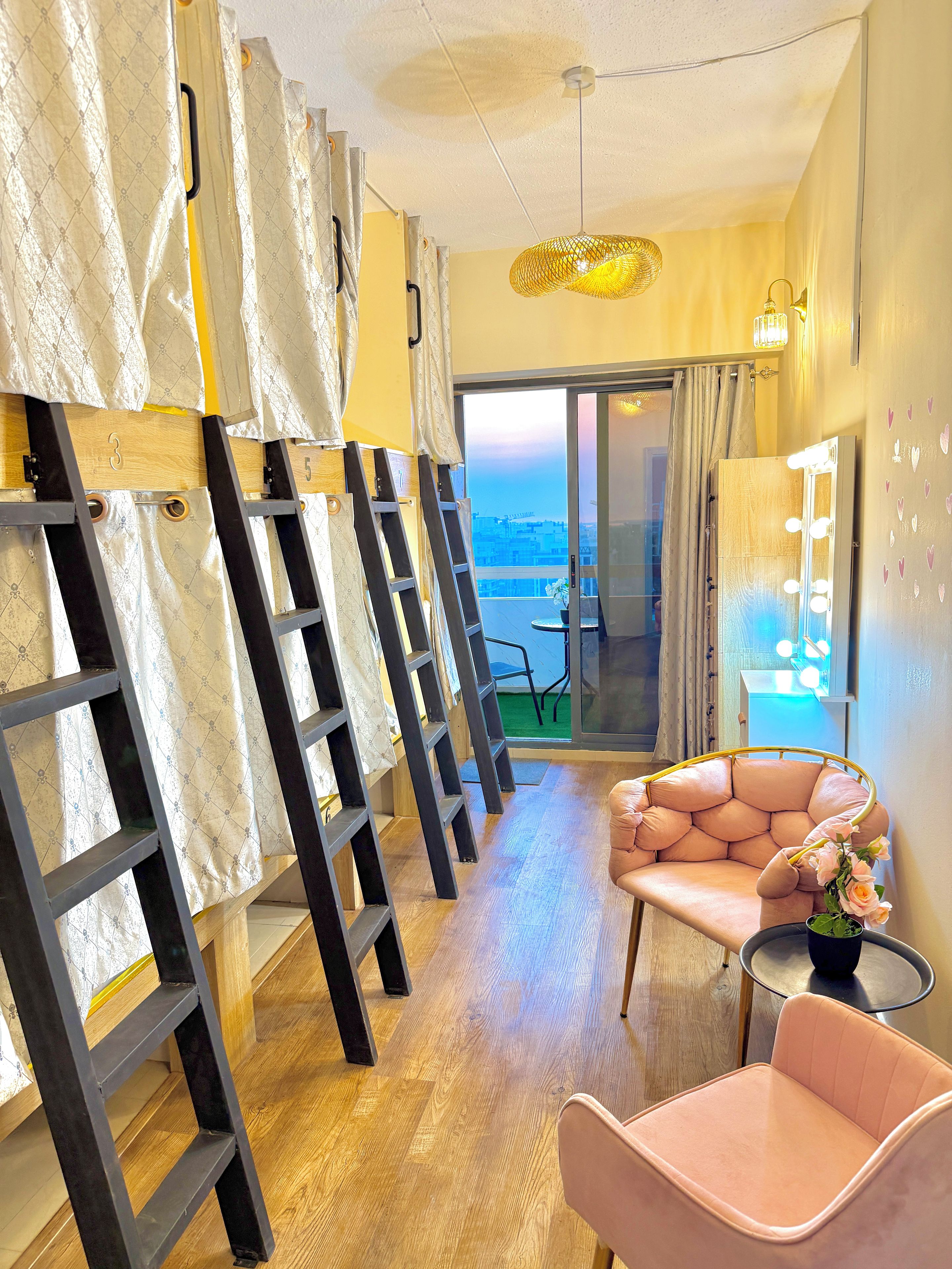A room with bunk beds, a vanity with a mirror and lights, a pink chair, and a view of the city.