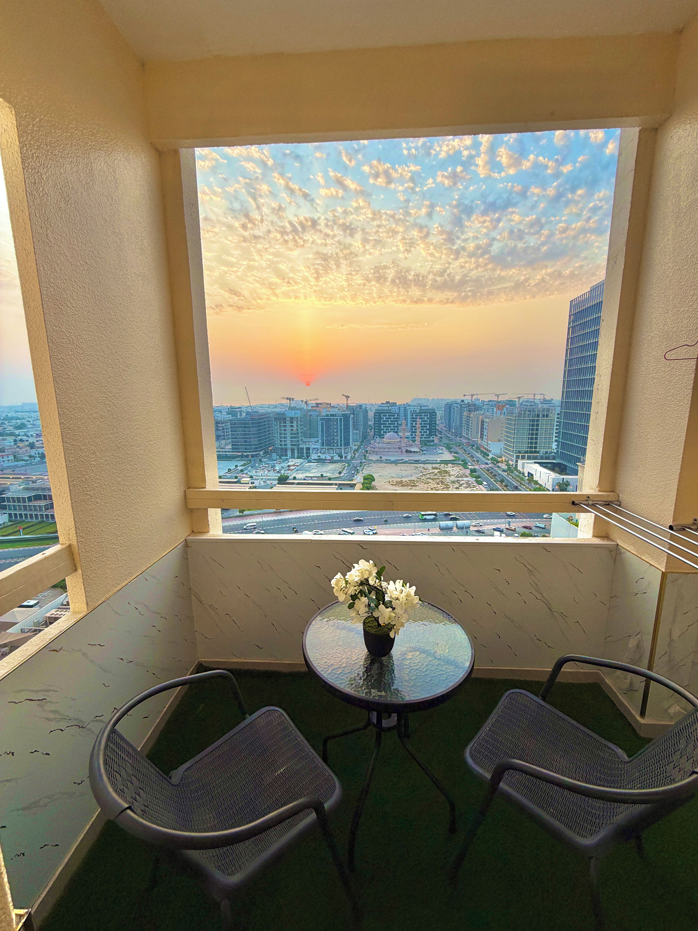 A balcony with a table and chairs, a vase of flowers, and a view of a cityscape at sunset.