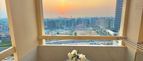 A balcony with a table and chairs, a vase of flowers, and a view of a cityscape at sunset.