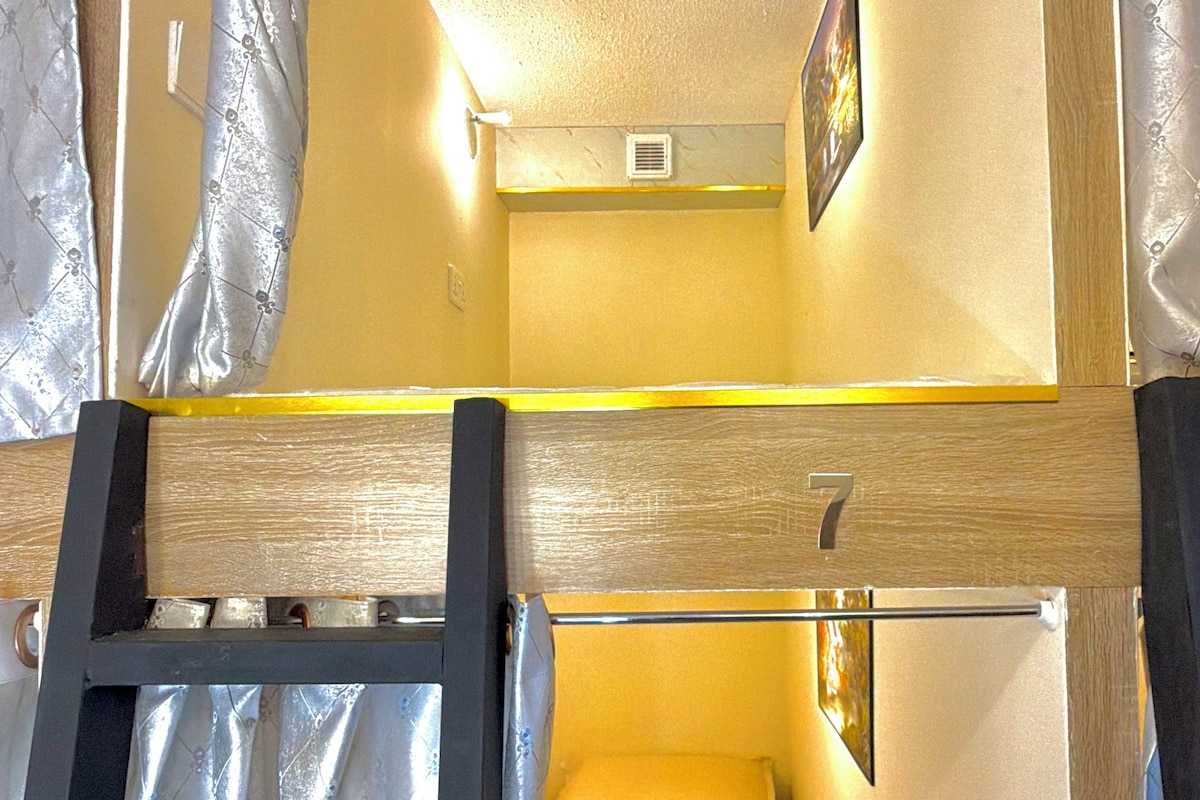 Un lit superposé avec une échelle, un lit simple, une petite fenêtre et un tableau accroché au mur.