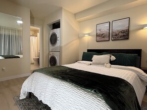 Een slaapkamer met een bed, een wasmachine, een spiegel en twee landschapsprenten aan de muur.