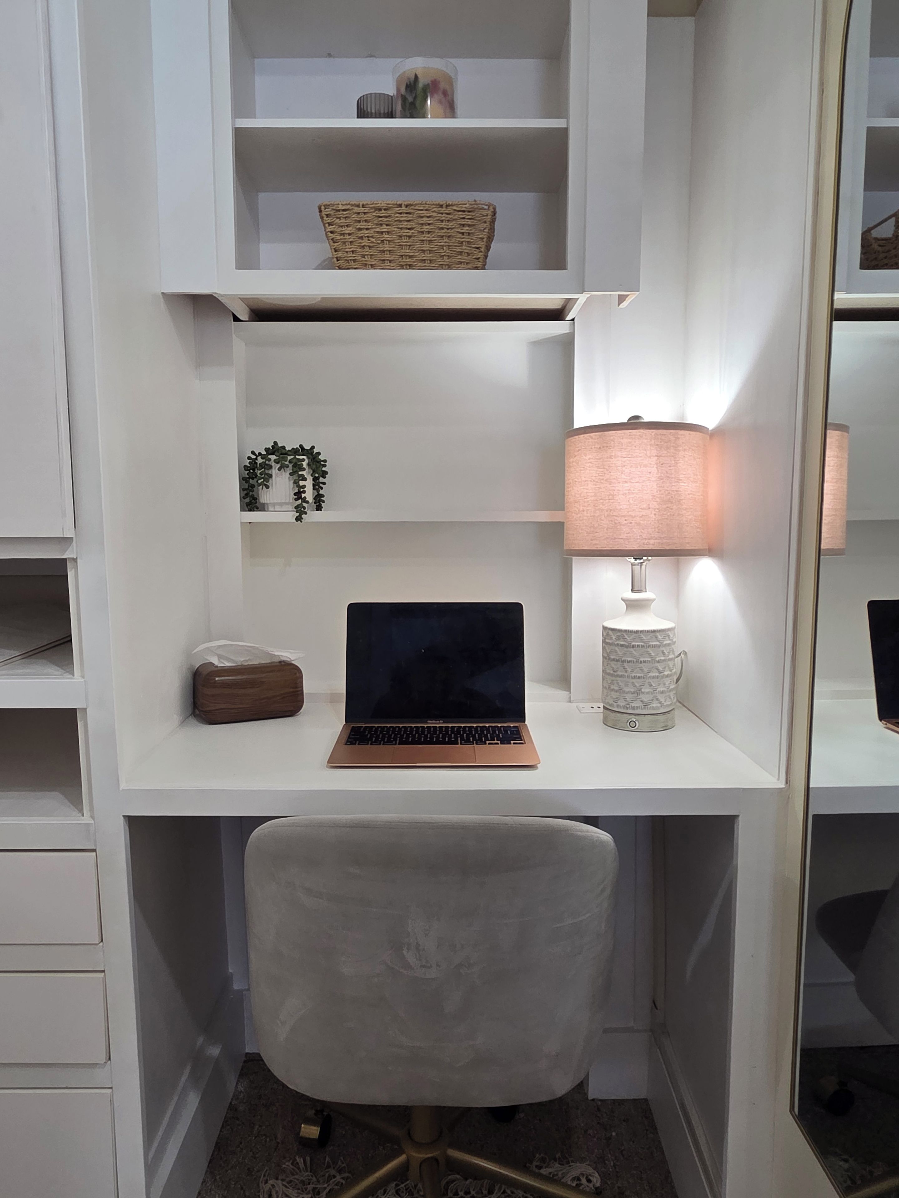 Un escritorio blanco con una computadora portátil, una lámpara, una cesta de mimbre y una planta pequeña.