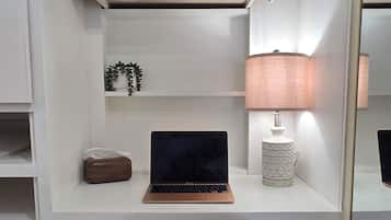 Un escritorio blanco con una computadora portátil, una lámpara, una cesta de mimbre y una planta pequeña.
