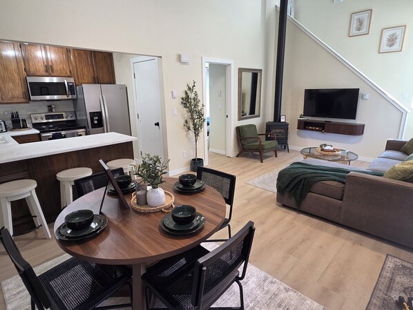Een moderne keuken met een magnetron, fornuis en koelkast. Er is een eethoek met een ronde tafel en stoelen, en een woonkamer met een bank en een televisie.