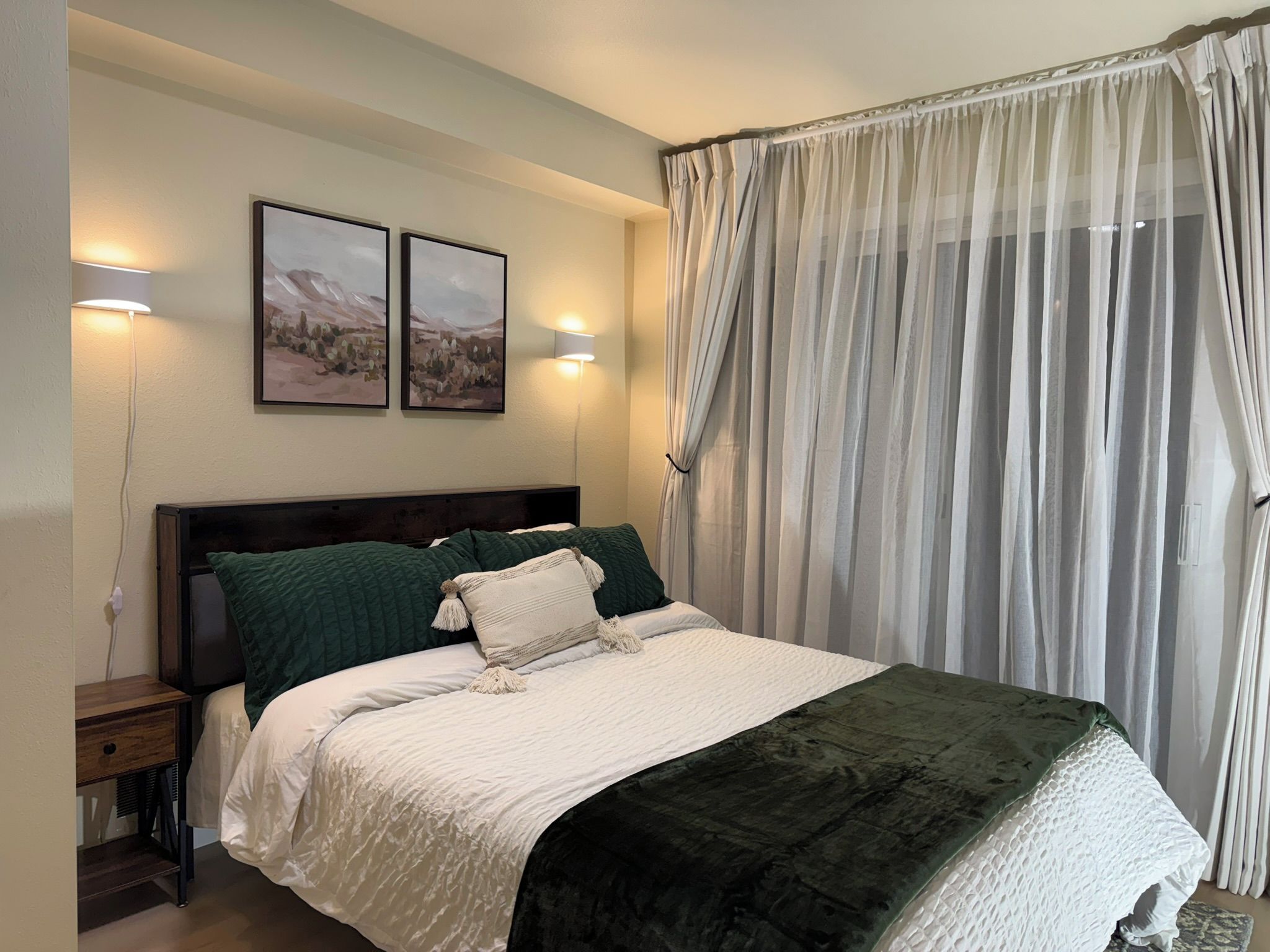 A bedroom with a bed, two wall-mounted lights, a nightstand, a dark wooden headboard, and a sheer curtain.
