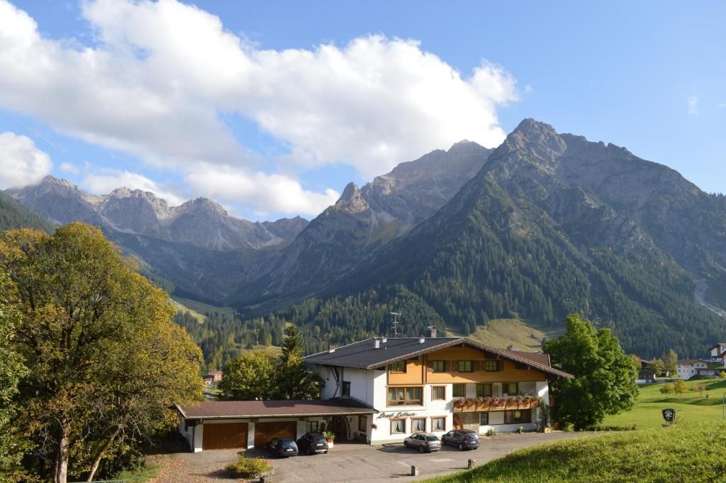 A hotel with a parking area, a building with a sloped roof, and mountains in the background.